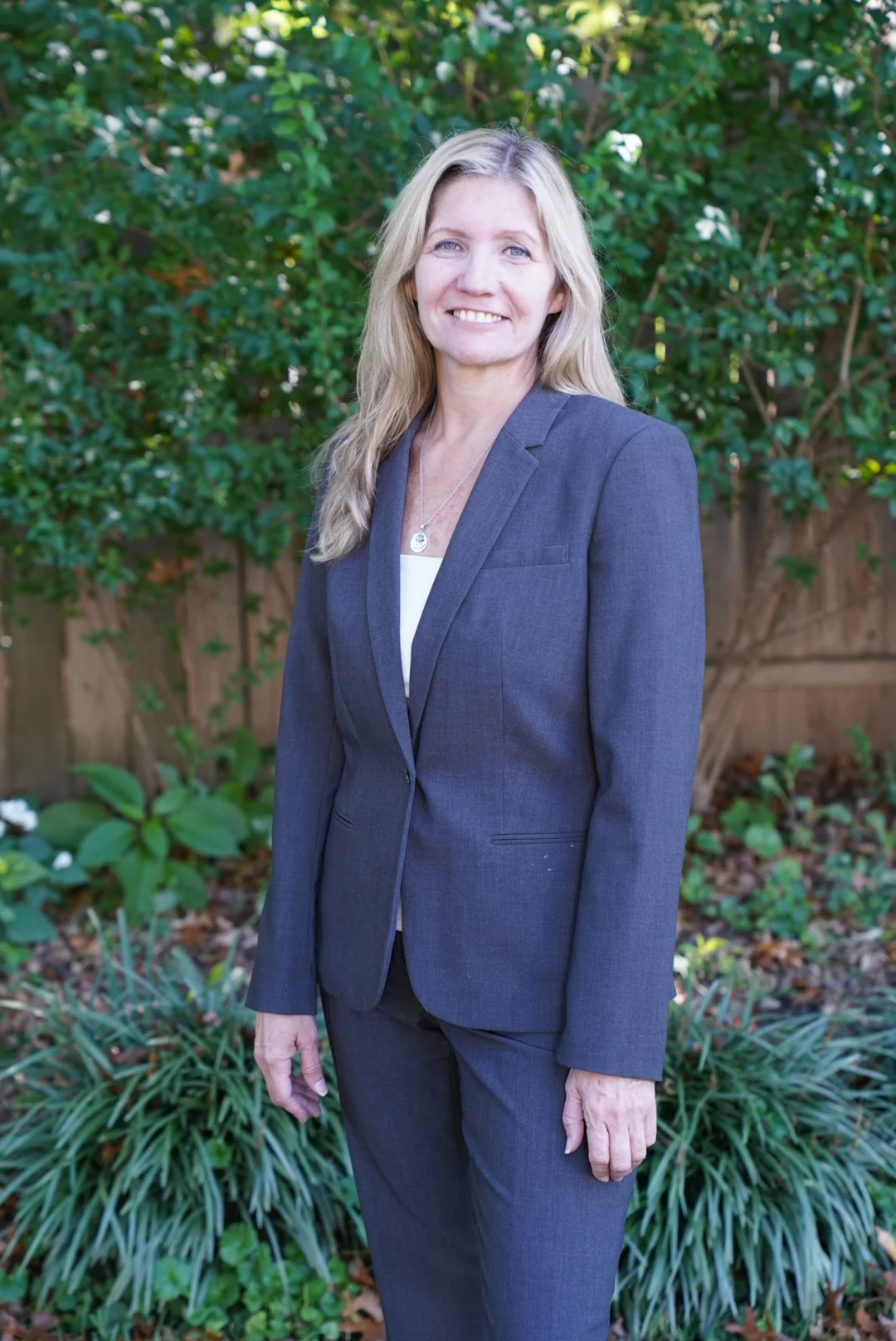Heather A. White smiling outdoors in a gray suit, with greenery behind her.